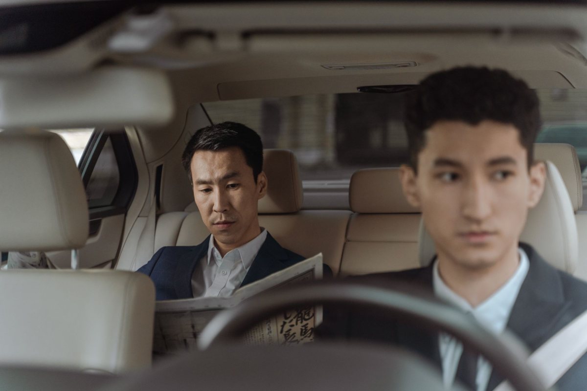 Chauffeur in a suit driving while a passenger reads a newspaper in the back seat